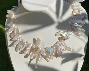 May include: A necklace composed of numerous irregular, elongated, iridescent white freshwater pearls. The pearls feature a high-luster finish with subtle pink and blue undertones, arranged in a single strand that drapes around the neck.