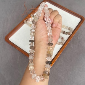 May include: A strand of faceted, clear, and smoky quartz beads with natural inclusions, strung on a thin cord. The beads vary in transparency and color, ranging from translucent white to dark brown, and are arranged in a continuous loop.