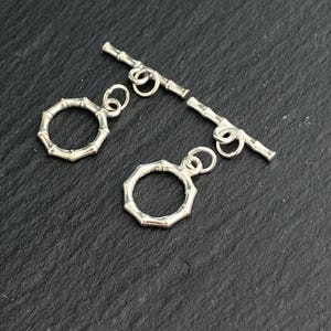 Puede incluir: Dos cierres de palanca de plata para joyería. Cada conjunto consta de un anillo circular con una textura segmentada similar al bambú y una barra en T a juego. Las piezas de metal tienen un acabado pulido y pequeñas anillas de unión.