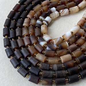 May include: A multi-strand necklace featuring cylindrical beads made of banded agate in shades of dark brown, tan, and cream. The beads are separated by small, metallic gold-toned spacer beads, creating a repeating pattern throughout the strands.