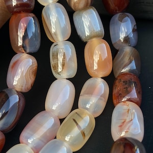 Puede incluir: Primer plano de un collar de cuentas de ágata pulidas en varios colores, incluyendo tonos de marrón, naranja y crema. Las cuentas de forma ovalada tienen un acabado brillante, con patrones naturales visibles. Las cuentas están ensartadas, listas para la fabricación de joyas.