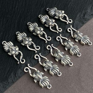 May include: A collection of nine silver-toned metal jewelry clasps, each shaped like a goldfish with detailed scales and fins. The clasps feature a hook-and-eye mechanism, with the fish head serving as the hook and the tail forming the eye loop.