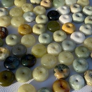 Pode incluir: Uma coleção de numerosas contas de pedra lisa e polida em forma de pequenos anéis ou roscas. As contas apresentam uma variedade de cores naturais, incluindo tons de verde, amarelo, branco, cinzento e preto, algumas com padrões marmoreados.
