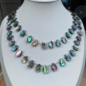 May include: A double-strand necklace featuring teardrop-shaped abalone shell beads. Each bead displays iridescent hues of green, blue, purple, and pink, strung on a clear cord. The beads are arranged in two rows, showcasing the natural shell texture.