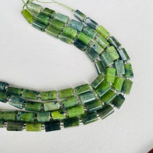 Pode incluir: Fios de contas retangulares verdes com espaçadores prateados. As contas variam em tons de verde, algumas com um padrão mosqueado. As contas são enfiadas em um cordão fino verde claro. As contas medem aproximadamente 1 cm de comprimento.