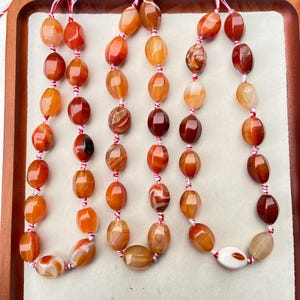 Può includere: Più fili di perline di pietre preziose nelle tonalità dell'arancione, del rosso e del marrone. Le perline ovali sono infilate su un cordoncino rosso e bianco. Le perline hanno una finitura lucida e variano per colore e motivo. Ogni perla misura circa 1,5 cm di lunghezza.