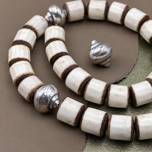 Peut inclure: Un collier composé de perles cylindriques blanc cassé aux extrémités brun foncé, alternées avec des perles en métal argenté poli. Une perle en métal argenté isolée avec une texture en spirale est placée près du collier enroulé.