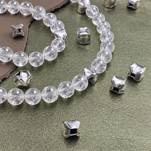 Può includere: Una collezione di perline rotonde trasparenti dall'aspetto crepato infilate in un motivo circolare, intervallate da piccole perline distanziatrici quadrate color argento metallizzato. Diverse perline quadrate color argento sono sparse.