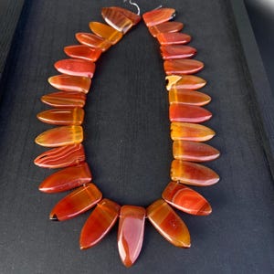 Puede incluir: Un collar compuesto por cuentas de ágata pulidas de color marrón rojizo. Las cuentas son alargadas y en forma de lágrima, con diferentes tonos de rojo, naranja y marrón. El collar se muestra sobre un fondo oscuro, resaltando los colores y patrones naturales de las piedras.