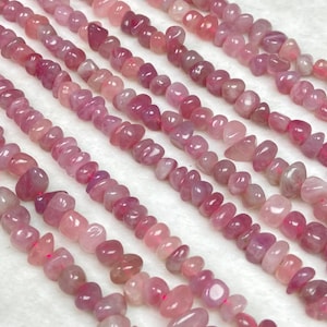 May include: Close-up of several strands of pink and rose-coloured gemstone beads. The beads are small, irregular in shape, and strung together. The colour varies from light pink to deep rose, creating a gradient effect. Likely used for jewellery making.
