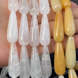 Può includere: Primo piano di fili di perline a forma di goccia in varie tonalità. Le perline variano dal bianco traslucido al giallo dorato. Ogni perlina è infilata, mostrando le loro superfici lucide e le variazioni naturali. Ideale per la creazione di gioielli.