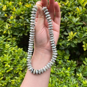 Può includere: Una collana di perline di pietre preziose bianche e grigie macchiate. Le perline di forma ovale sono infilate su un sottile cordino trasparente. Lo sfondo è un cespuglio verde.