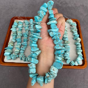 Può includere: Una collezione di perline in pietra turchese infilate su un filo sottile. Le perline hanno forme irregolari e levigate in un colore blu ciano brillante con venature di matrice marrone visibili. I fili sono disposti su un vassoio rettangolare.