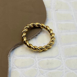 Op de afbeelding: Een goudkleurige ring met een doorlopend gedraaid touwontwerp. Het metalen oppervlak is gepolijst en reflecterend, waardoor een reeks onderling verbonden diagonale ribbels ontstaat die de cirkelvormige band van het sieraad vormen.