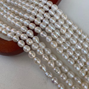Puede incluir: Cadenas de perlas blancas de agua dulce y brillantes. Las perlas tienen forma de lágrima y un brillo iridiscente sutil. Las perlas están ensartadas, listas para la fabricación de joyas. Las perlas se muestran sobre una superficie de madera.