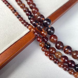 May include: Close-up of several strands of reddish-brown gemstone beads. The beads vary in size and shade, with some appearing nearly black. The beads are displayed on a wooden surface and a white background.
