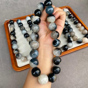 May include: A strand of round, polished beads featuring a mix of solid black, translucent grey, and banded agate patterns. The beads are strung together in a continuous line, showcasing variations in colour, opacity, and natural stone textures.