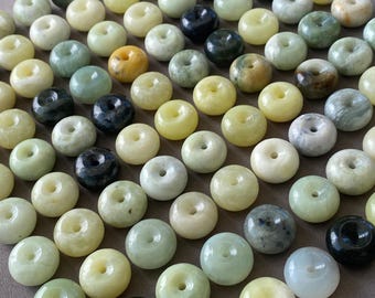 May include: A collection of numerous smooth, round stone beads, each featuring a central hole. The beads display a variety of natural colors, including shades of pale green, cream, yellow, dark gray, and black, arranged in a grid-like pattern.