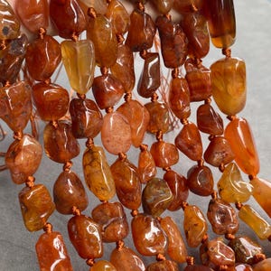 Peut inclure: Une collection de perles en pierre de cornaline polies et irrégulières dans des tons orange brûlé, ambre profond et brun rougeâtre. Chaque pierre est nouée individuellement sur un fin cordon de couleur cuivre, formant plusieurs brins verticaux.