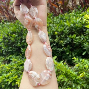 May include: A necklace with large, polished agate stones in pink, peach, and white. The stones are irregularly shaped and strung on a pink cord. The necklace is a striking accessory. The background is green foliage. The necklace is a unique piece of jewellery.