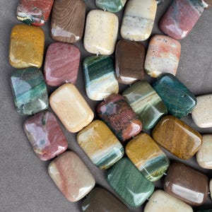 May include: A collection of polished, rectangular stone beads featuring a variety of natural patterns and colours, including shades of green, brown, yellow, red, and cream. Each bead has a smooth, glossy finish and rounded corners.