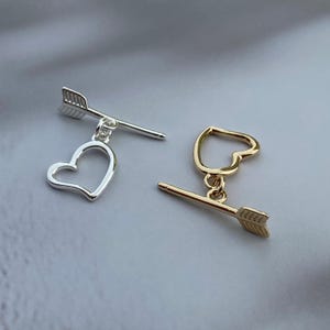 Puede incluir: Dos cierres de palanca para joyería. Un cierre es de tono plateado con una barra recta en forma de flecha y un bucle en forma de corazón. El segundo cierre es de tono dorado, con una barra de flecha a juego y un bucle en forma de corazón.