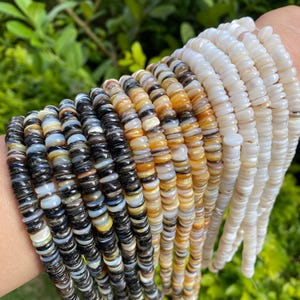 May include: Several beaded necklaces in shades of black, tan, and white. The small, flat beads are strung together, creating a layered look. The necklaces are shown against a natural green backdrop.