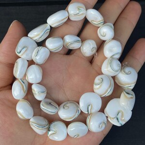 Può includere: Una collana di perline bianche e color crema con motivi a vortice beige e verdi. Le perline sono di varie forme e dimensioni, infilate insieme. Le perline sono tenute in una mano.