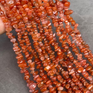 Peut inclure: Plusieurs brins de petites pépites de cornaline polies et irrégulières. Les pierres présentent une gamme de teintes orange, orange rougeâtre et rouge brunâtre, allant du translucide à l'opaque, avec une texture lisse et polie.