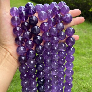Op de afbeelding: Een verzameling gladde, ronde amethistkralen gerangschikt in meerdere verticale strengen. De kralen vertonen verschillende tinten paars, variërend van licht lavendel tot diep violet, met natuurlijke variaties in kleur en doorzichtigheid.