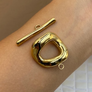Può includere: Un set di chiusura a T color oro composto da un componente a barra allungata e liscia con un occhiello centrale e un componente ad anello triangolare arrotondato di forma irregolare con un piccolo occhiello di fissaggio sul bordo inferiore.