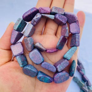 May include: A strand of gemstone beads in shades of purple and teal. The beads are rectangular with a cracked, textured appearance. The beads are strung together, ready for jewelry making.
