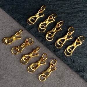 May include: A collection of eight gold-toned metal jewelry clasps, each featuring a hook-and-eye design with a coiled wire detail at the center. These small fasteners are arranged in a scattered pattern, showcasing a polished metallic finish.