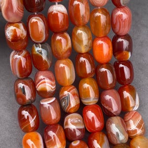 Può includere: Una collezione di perline di corniola levigate a forma di barilotto, disposte in fili verticali. Le perline mostrano una gamma di colori caldi tra cui rosso intenso, arancione bruciato e ambra traslucida, con motivi naturali a bande.