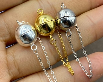 May include: Three spherical magnetic jewelry clasps, one in silver tone, one in gold tone, and one in dark gunmetal tone. Each smooth, round clasp features a delicate metal chain attached to the bottom, designed for securing necklaces or bracelets.