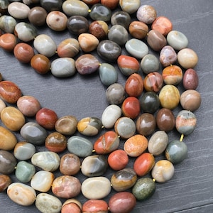 Puede incluir: Un collar de piedras de río pulidas, lisas y de forma ovalada en una variedad de colores tierra que incluyen tonos marrón, gris, beige, óxido y crema. Las piedras están ensartadas en un cordón amarillo fino, formando un bucle continuo.