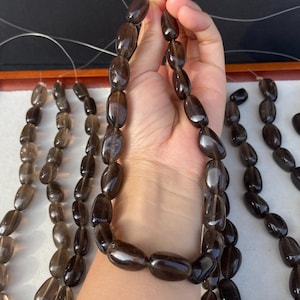 Puede incluir: Un collar de cuentas de cuarzo ahumado pulidas, de forma ovalada. Las cuentas son de color marrón translúcido y están ensartadas. Se ven varias otras hebras de cuentas similares en el fondo.