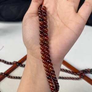 May include: A strand of small, round, reddish-brown beads. The beads are strung together, forming a necklace or bracelet. The beads are held in a hand, with a wooden structure in the background.