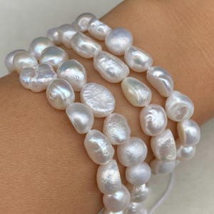 Op de afbeelding: Drie strengen witte, onregelmatig gevormde parels. De parels hebben een glanzende, iriserende glans. De strengen zijn naast elkaar gerangschikt, wat wijst op een armband of ketting. De parels zijn geregen op een wit koord.