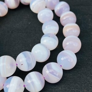 Pode incluir: Close-up de um fio de contas redondas listradas em rosa claro e branco. As contas são enfiadas com um fio roxo. As contas têm um acabamento polido e brilhante e estão dispostas em uma linha curva.