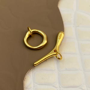 Puede incluir: Un conjunto de cierre de palanca en tono dorado que consta de un anillo circular y un componente en forma de T. La barra en T presenta el texto grabado S925. Ambas piezas tienen un acabado metálico liso con formas orgánicas irregulares.