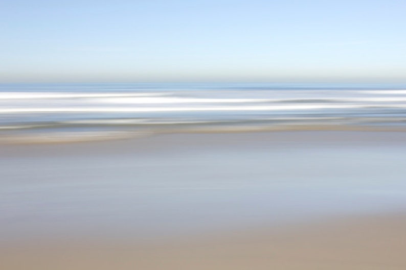 May include: A blurred image of a beach with white waves crashing on the shore. The sky is a light blue and the sand is a light brown.