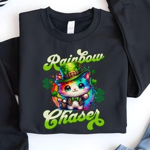Puede incluir: Sudadera negra con un gato arcoíris que lleva un sombrero verde y sostiene un pote de oro. El texto "Rainbow Chaser" está escrito en verde y blanco.