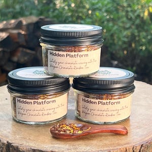 May include: Three glass jars with black lids containing loose leaf tea. The jars are labeled "Hidden Platform" and the text "Satisfy your chocolate craving with the Belgian Chocolate Rooibos Tea".