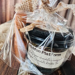 May include: A gift set featuring a jar of Belgian Chocolate Rooibos Tea, packaged in clear cellophane with a burlap ribbon. The jar has a black lid and a label that reads "Hidden Platform". A wooden spoon is included.