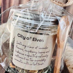 Puede incluir: Un tarro de cristal de té etiquetado "City of Elves" con una cuchara de madera. El tarro contiene una mezcla de flores de lavanda, hojas de frambuesa e hibisco. El té está empaquetado en celofán transparente y atado con una cinta de arpillera.