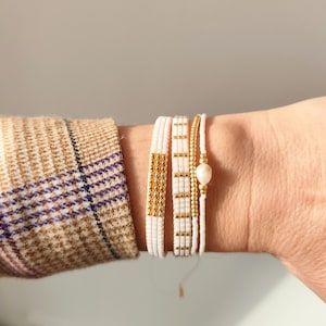 Peut inclure: Quatre bracelets perlés blancs et dorés. L'un des bracelets présente un motif de perles dorées, tandis que les autres ont des accents dorés. Un seul bracelet a une perle. Les bracelets sont sur un poignet, avec une manche de veste à carreaux en arrière-plan.