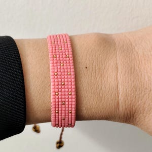 May include: Pink beaded bracelet with gold-colored accents. The bracelet is made of small, evenly spaced beads, creating a textured surface. The bracelet is on a wrist, and the background is a plain white surface.