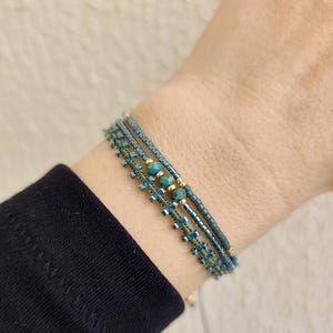 Peut inclure: Trois bracelets de perles en tons de teal et d'or sont empilés sur un poignet. Les bracelets présentent de petites perles et des éléments décoratifs. Le poignet est couvert par un haut à manches longues noir.