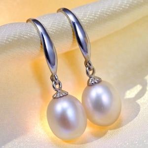 Op de afbeelding: Een paar elegante parel oorbellen. Elke oorbel heeft een zilveren haak en een grote, ovale, crèmewitte parel. De oorbellen worden gepresenteerd op een zachte, lichtgele stof, wat een luxueus gevoel creëert. De parels hebben een subtiele glans.